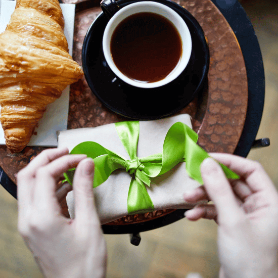 Two hands finish a bowtie wrapped gift. The gift sits on a table beside a cup of coffee and croissant.