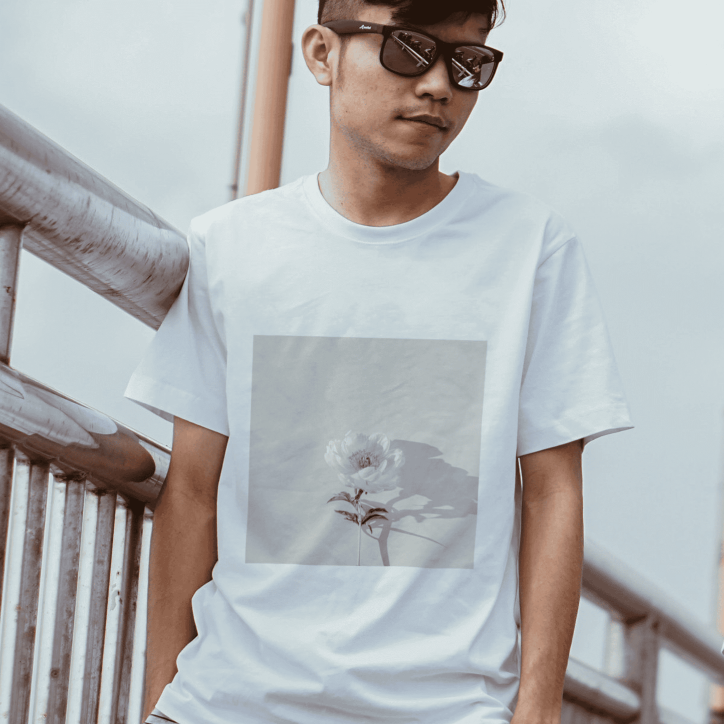 Young man wearing a white t-shirt featuring a delicate flower design, leaning against a railing.