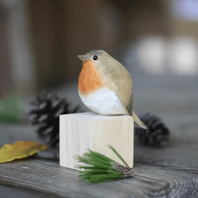 Hand carved bird ornament sitting on a wooden block with pinecones and leaves in the background.