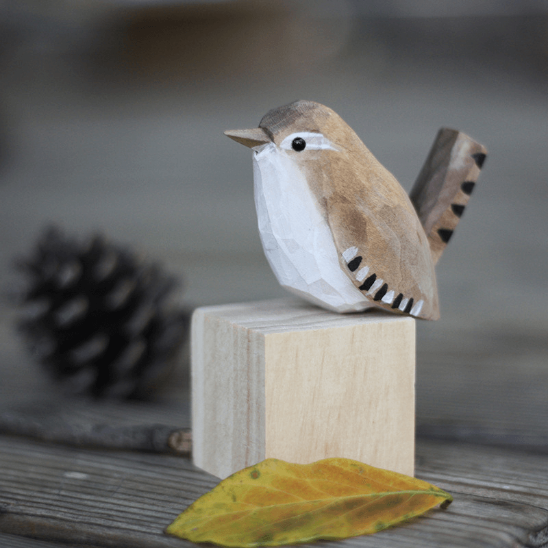 Hand-carved wooden bird ornament perched on a block with a pinecone and autumn leaf background.