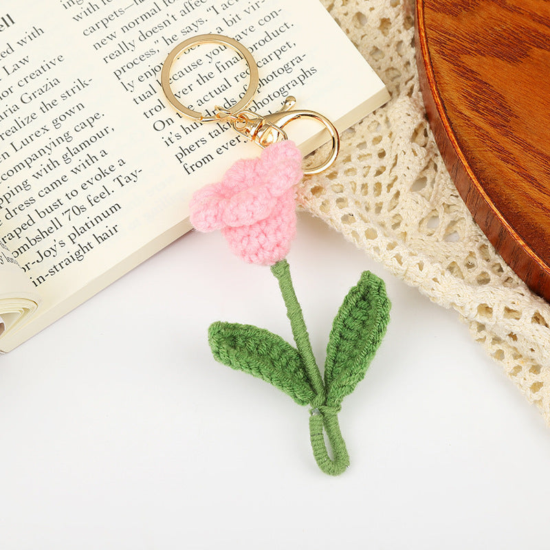 Crochet flower keychain in pink with green stem, placed on a book and lace, a unique handmade gift under $20.