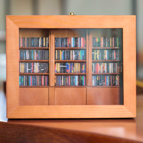 The Original Anxiety Bookshelf sits on a wood table. The mini books are all organized with the spines facing out. This replicates a miniature library in a box.