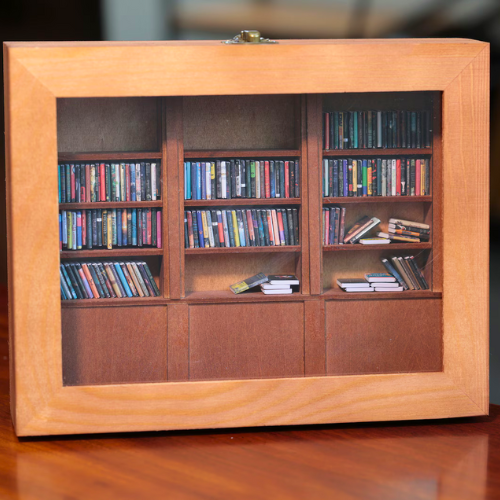 The Original Anxiety Bookshelf mini bookcase sits on a luxurious table. The mini bookshelf is filled with tiny books that are organized and shaken.