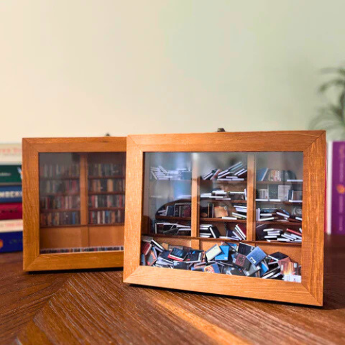 Two Anxiety Bookshelves sit on a table filled with miniature books. The mini bookshelf is filled with miniature replica books. It is a shakeable book box that you organize, stack, and shake to reduce stress and anxiety.