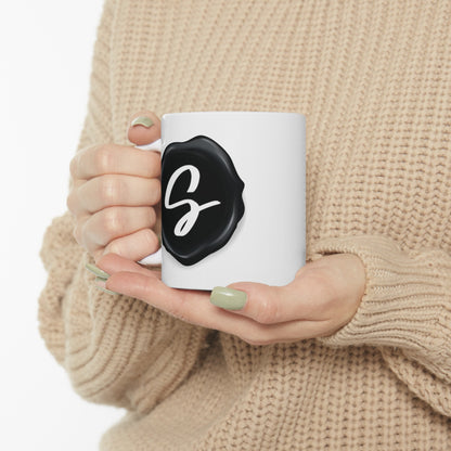 Sydney's Collection black wax seal logo is featured on a coffee mug. Held by a woman in a sweatshirt.