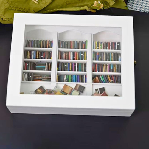 White Anxiety Bookshelf with books displayed as desk decor or bedroom decor. Some of the mini books are organized on the shelves, and some are disorganized on the tabletop. The white anxiety bookshelf is a miniature bookcase in a box with a door that opens to access the included miniature books.