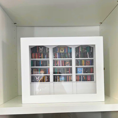 White Anxiety Bookshelf with books displayed as desk decor or bedroom decor. Some of the mini books are organized on the shelves, and some are disorganized on the tabletop. The white anxiety bookshelf is a miniature bookcase in a box with a door that opens to access the included miniature books.